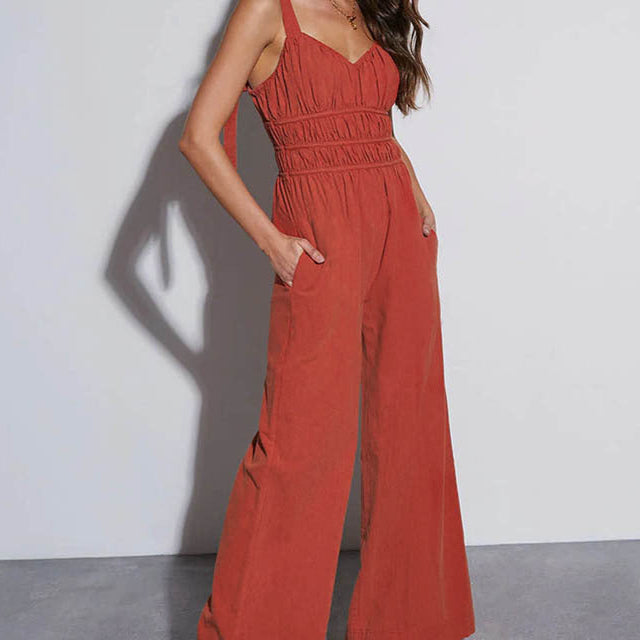 A woman in a rust-tone jumpsuit with ruched detailing and wide legs, accessorized with heels and jewelry.
