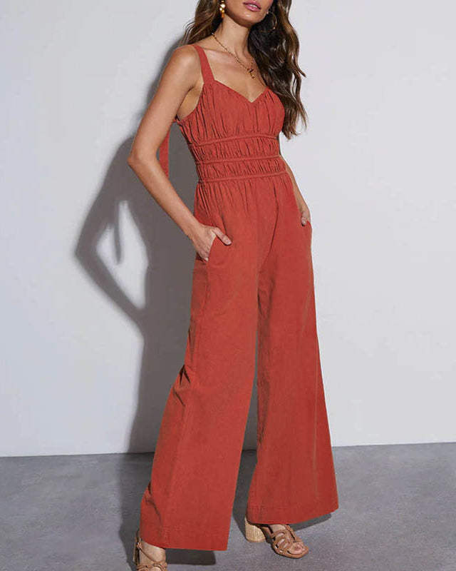 A woman in a rust-tone jumpsuit with ruched detailing and wide legs, accessorized with heels and jewelry.

