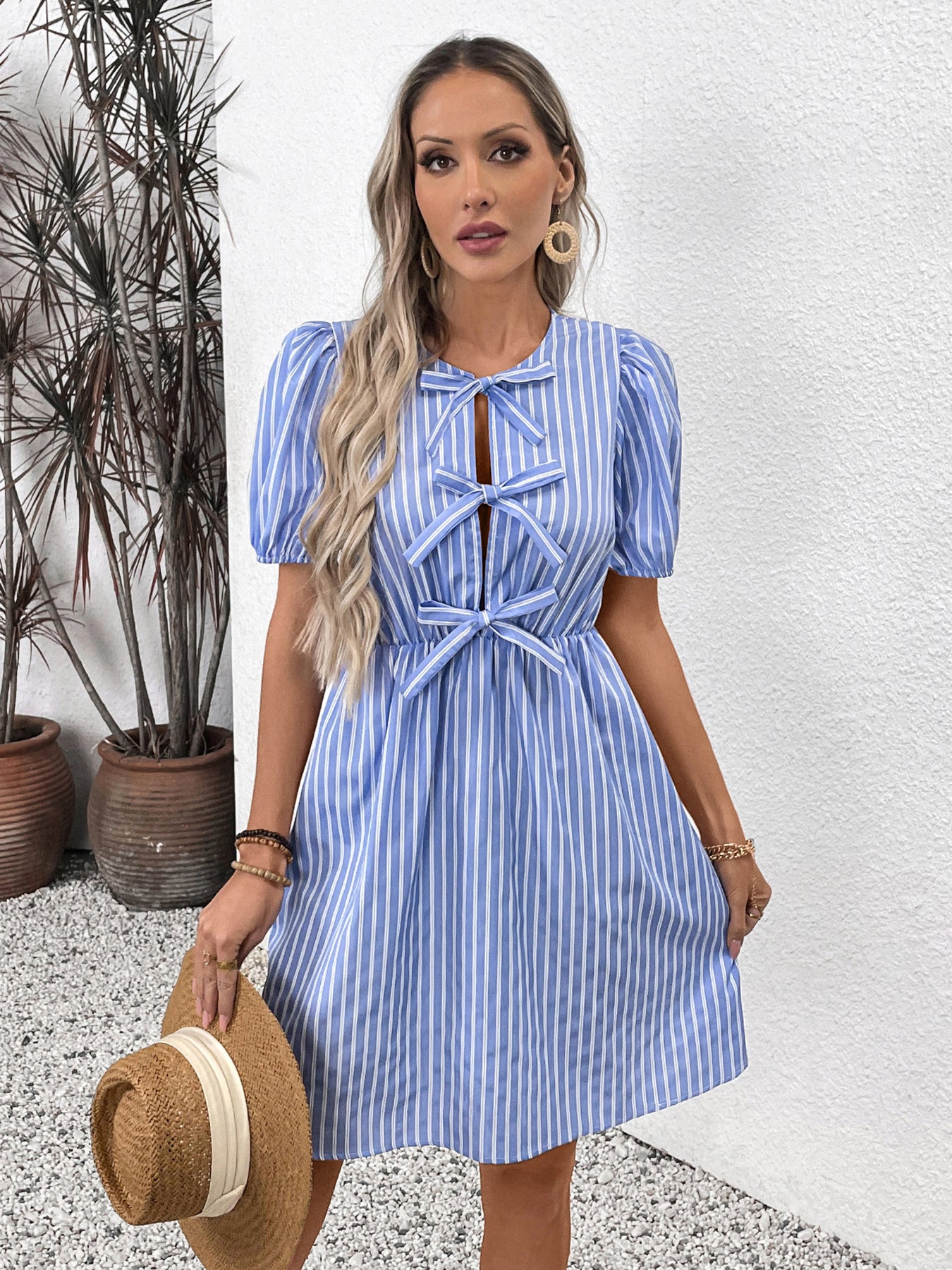 A model in a blue striped dress holds a straw hat while posing against a white background.