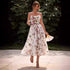 A model is holding a champagne glass and fan while wearing a white floral midi dress with pink roses, walking on a patio with garden background