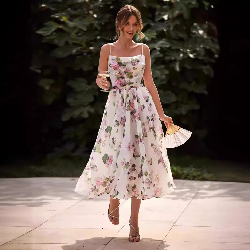 A model is holding a champagne glass and fan while wearing a white floral midi dress with pink roses, walking on a patio with garden background