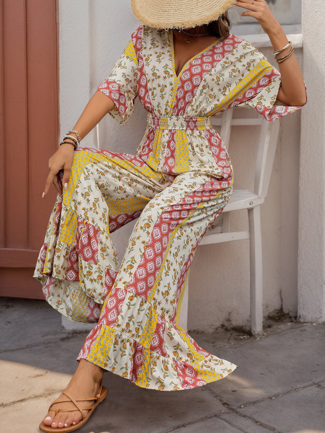 Woman sitting in a boho jumpsuit with a straw hat, featuring a deep V-neck, floral patchwork, and flowy wide-leg pants.