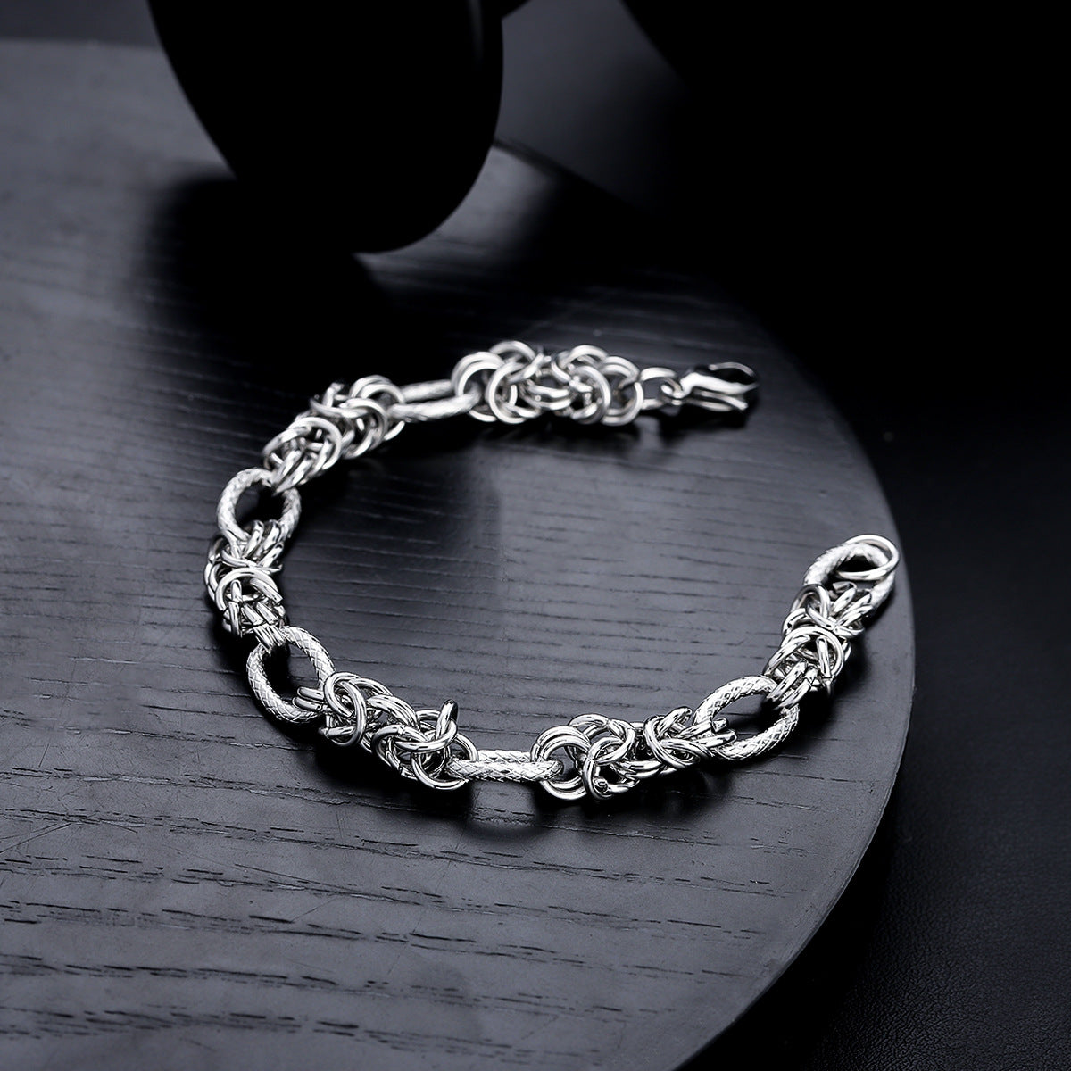 A silver rope-style chain bracelet placed on a black circular plate, showing its intricate knot design and polished finish.