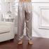 Model wearing gray geometric patterned silk pajama pants with light border at hem, standing barefoot on wooden floor near white furniture