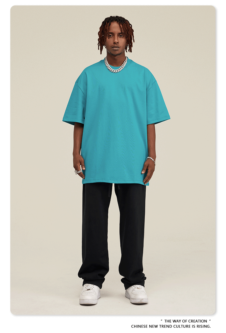 Man with short dreadlocks wearing turquoise oversized t-shirt, black sweatpants, white sneakers and silver chain necklace against neutral background