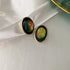 Close-up of oval iridescent green stud earrings with gold rim settings displayed on textured white background showing detailed craftsmanship