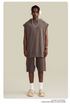 A model wearing taupe sleeveless hoodie with matching shorts, white headphones, silver chain necklace and cream slides against beige backdrop