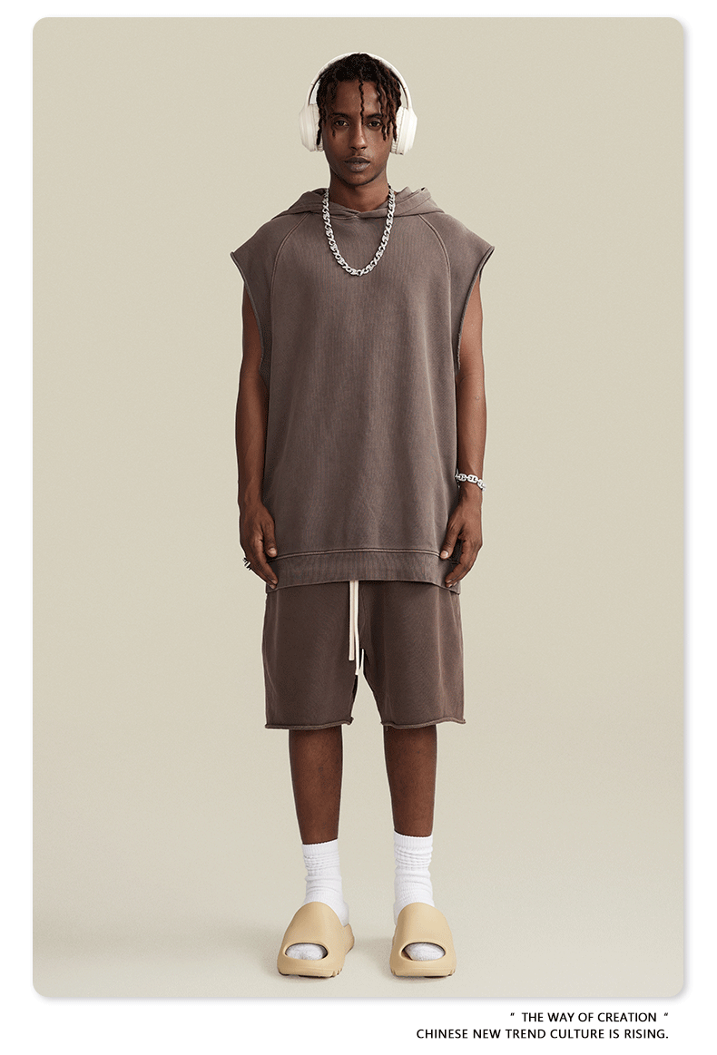 A model wearing taupe sleeveless hoodie with matching shorts, white headphones, silver chain necklace and cream slides against beige backdrop