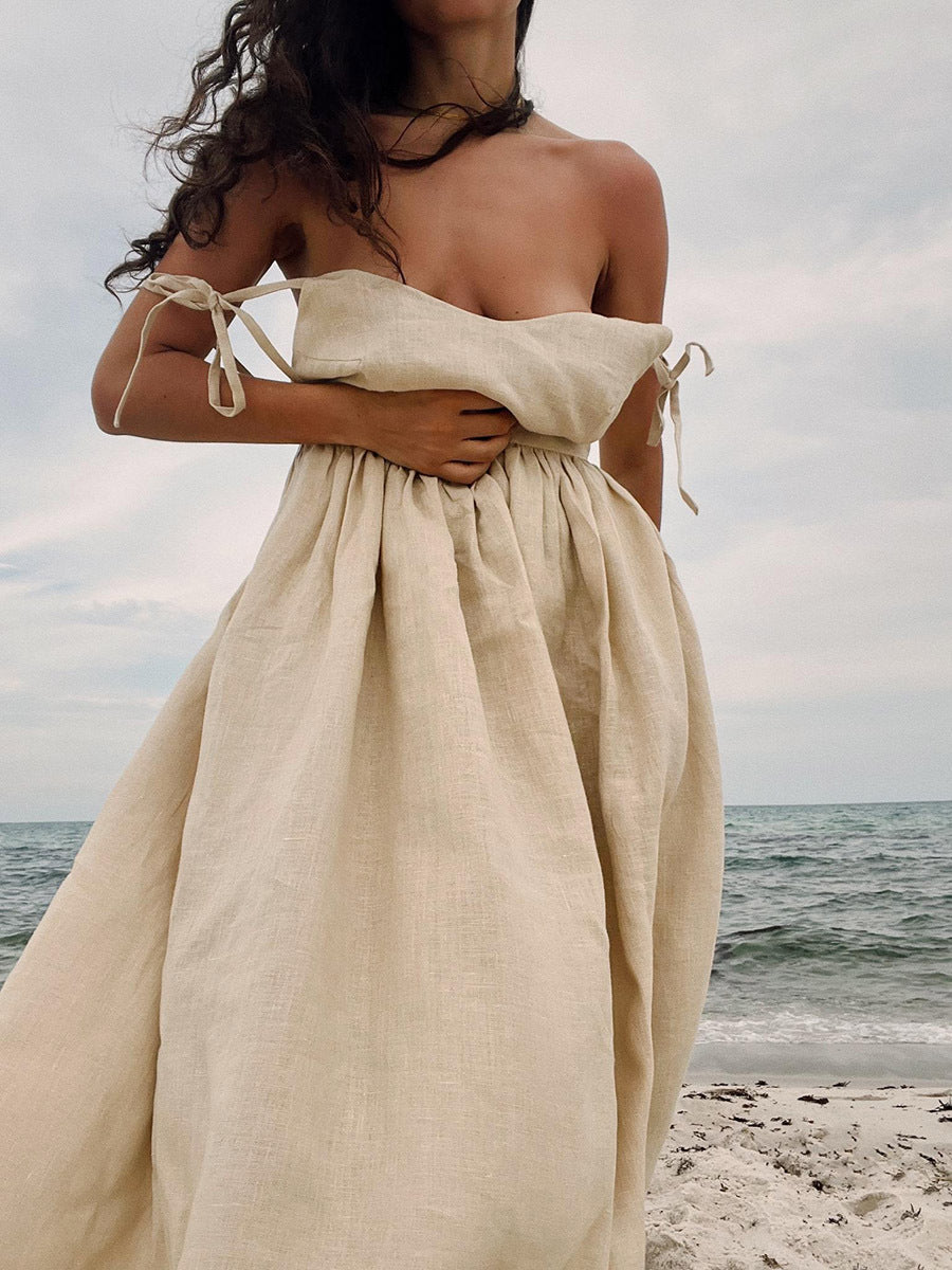 A model is wearing a natural beige linen tie-shoulder dress with gathered waist, standing at the beach with ocean backdrop