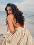 A model with curly hair is looking over her shoulder wearing a cream linen dress at the beach with ocean waves visible
