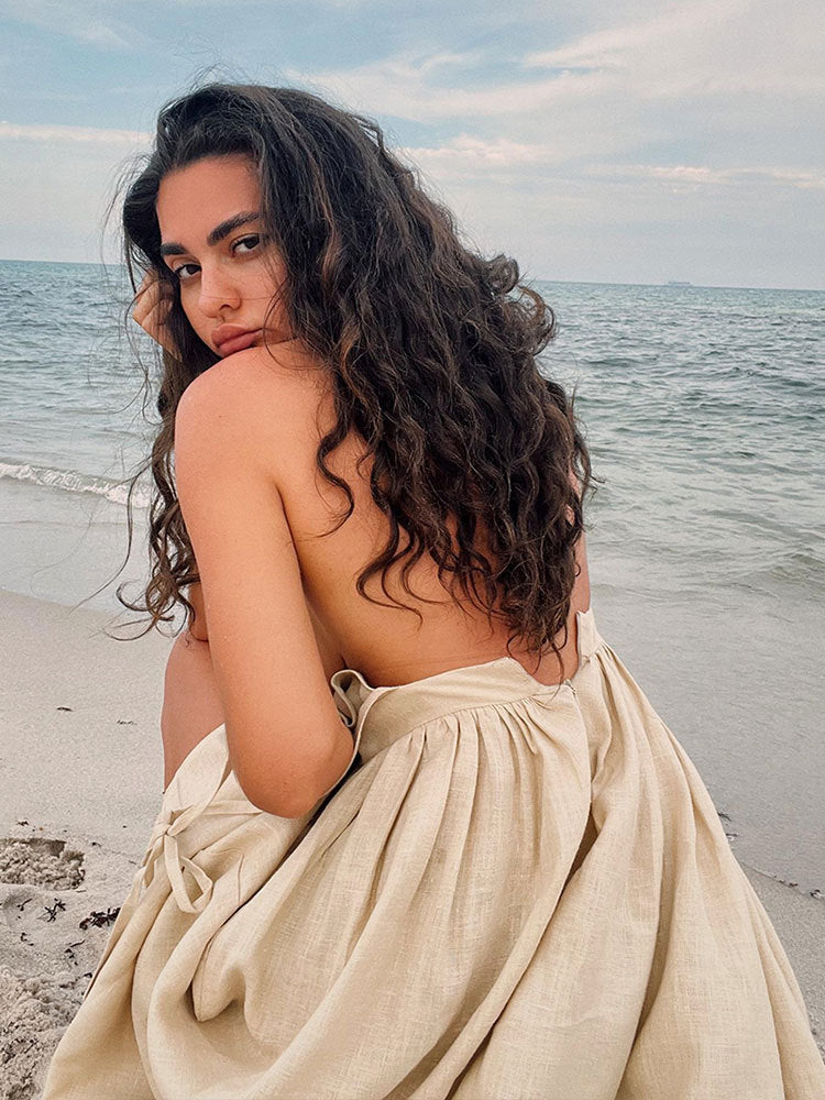 A model with curly hair is looking over her shoulder wearing a cream linen dress at the beach with ocean waves visible