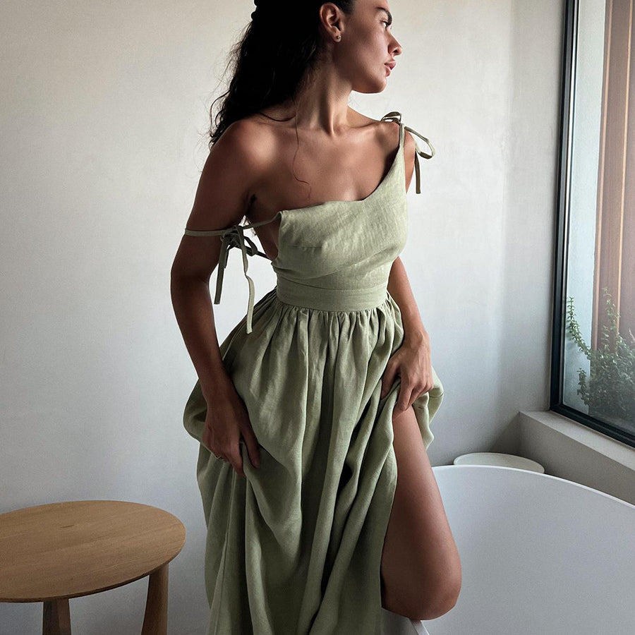 A model is wearing a sage green linen tie-shoulder dress with gathered skirt, standing in profile by a window with natural light