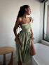 A model is wearing a sage green linen tie-shoulder dress with gathered skirt, standing in profile by a window with natural light