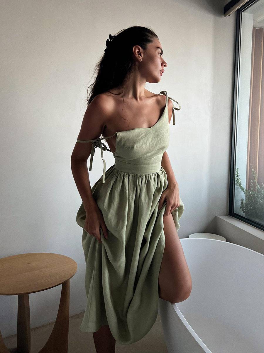 A model is wearing a sage green linen tie-shoulder dress with gathered skirt, standing in profile by a window with natural light