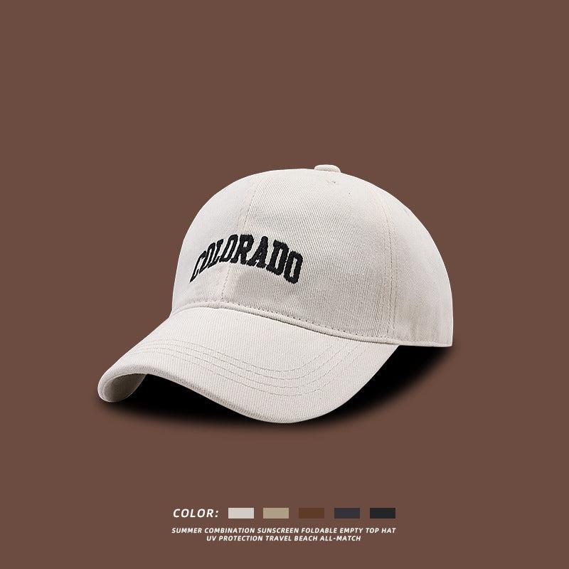 White baseball cap with navy COLORADO embroidery displayed against brown background, featuring UV protection and foldable design