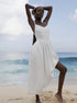 A model is standing on beach sand wearing a white linen sundress with hands behind head against ocean backdrop