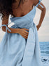 A model wearing a light blue linen tie-shoulder dress with gathered waist, holding fabric against ocean background