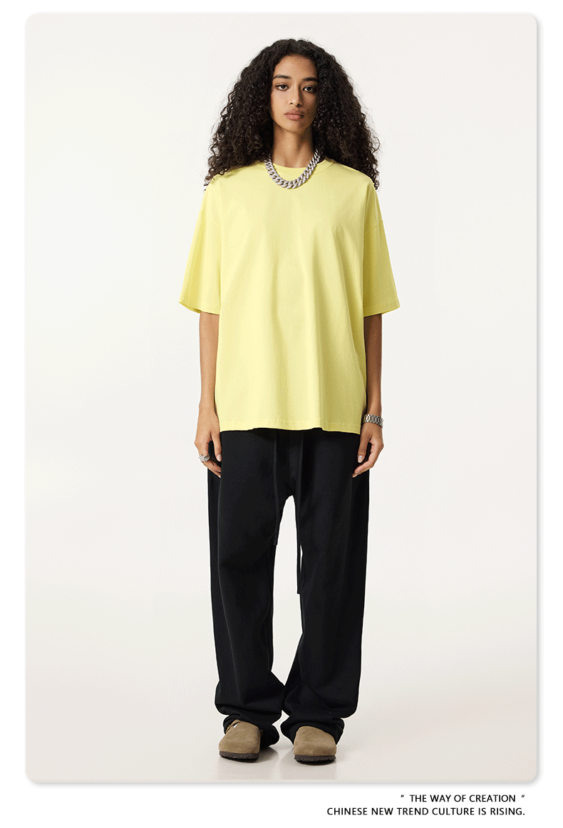 Female model wearing pale yellow oversized t-shirt with black wide-leg pants, styled with silver chain necklace and beige footwear against white background