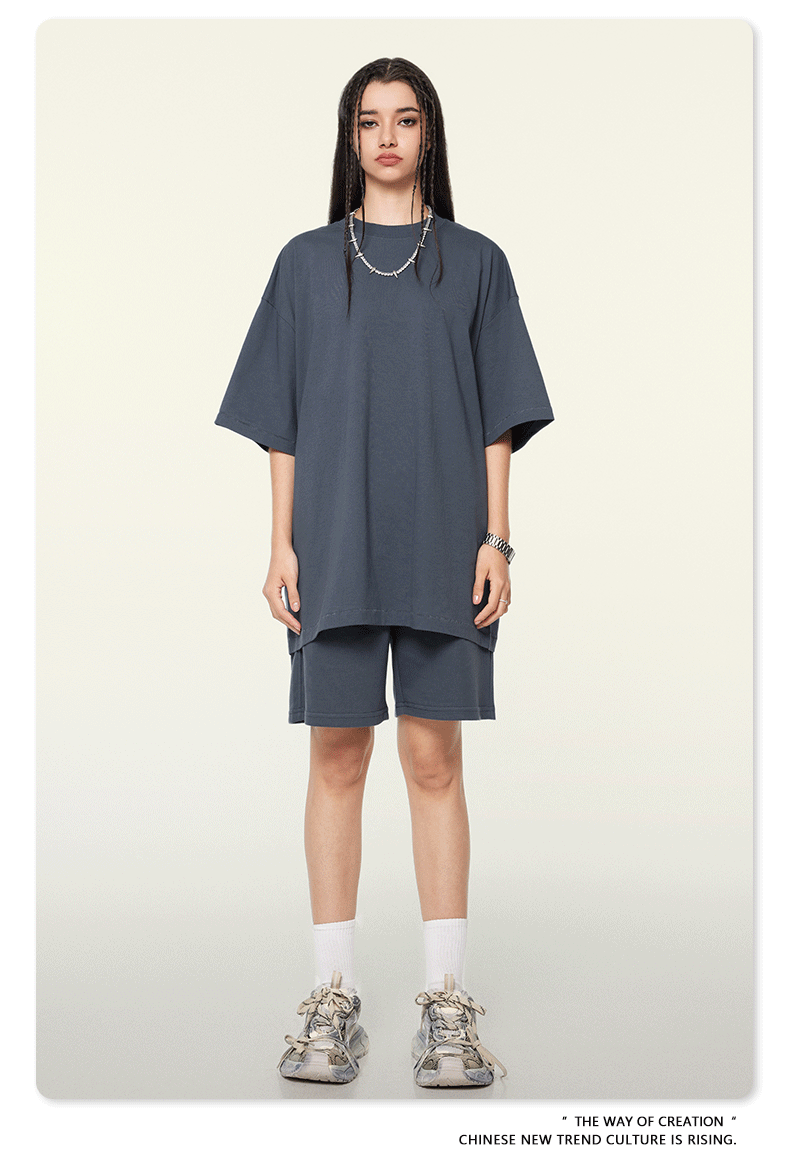 A woman wearing a charcoal gray oversized t-shirt and shorts set, accessorized with silver necklace, white socks, and chunky sneakers against neutral background