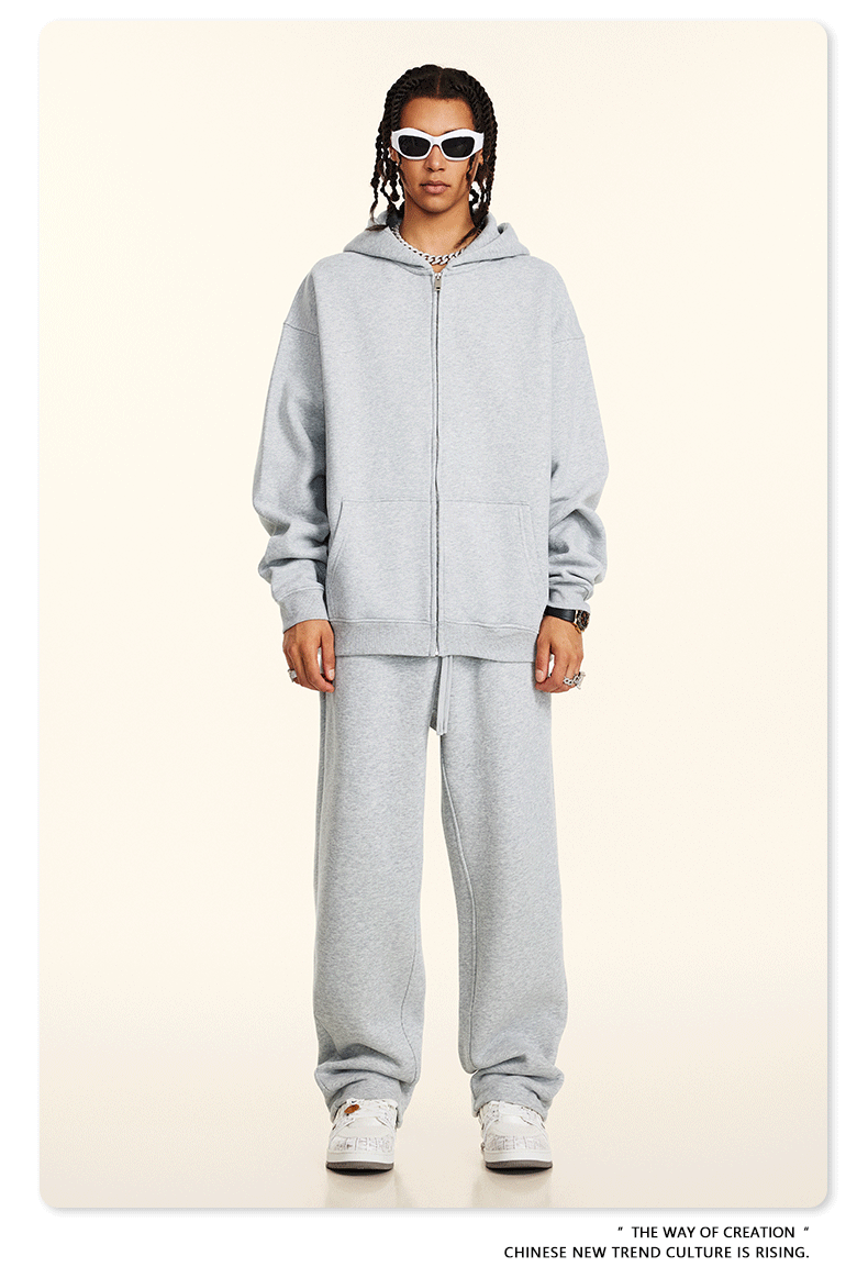 Model wearing light gray oversized zip-up hoodie and matching sweatpants with white rectangular sunglasses and silver chain necklaces against cream background