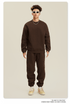 Male model in chocolate brown sweatsuit with crew neck sweatshirt and matching joggers, wearing white-framed sunglasses and beige sneakers