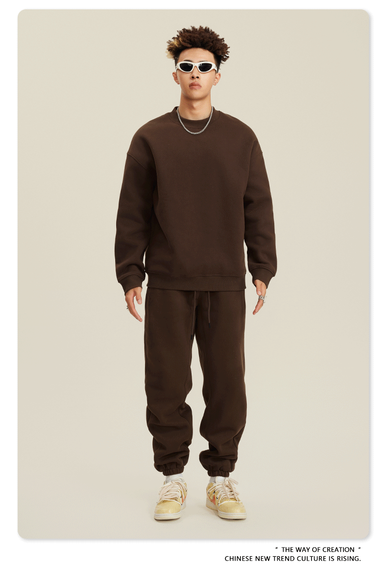 Male model in chocolate brown sweatsuit with crew neck sweatshirt and matching joggers, wearing white-framed sunglasses and beige sneakers