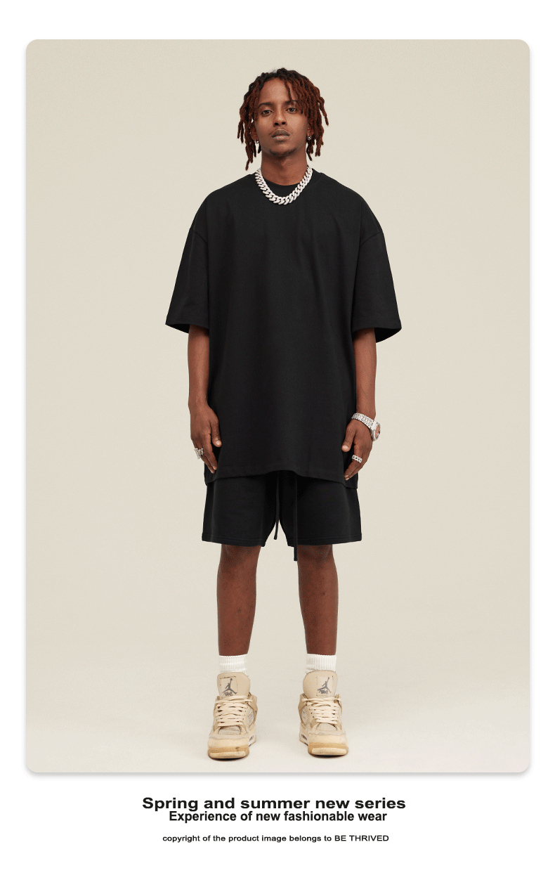 Model with dreadlocks wearing black oversized t-shirt and matching shorts with silver chain necklace and cream sneakers against cream background