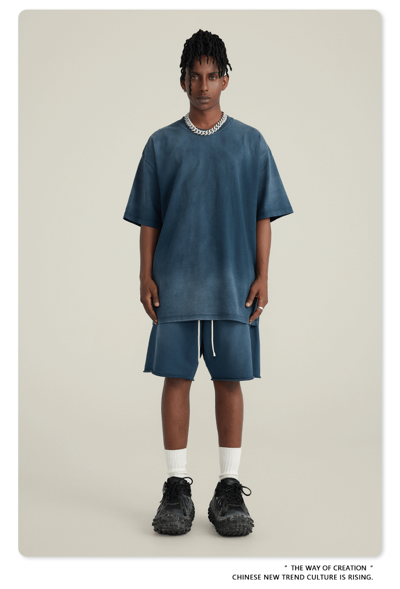 Model with short locs wearing navy blue t-shirt and shorts set with silver chain necklace, white socks and black sneakers against cream background