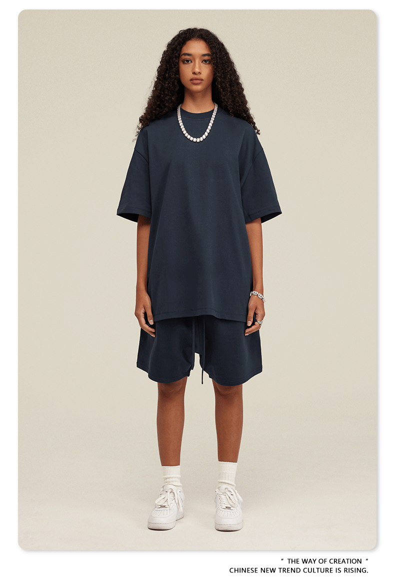 A woman with curly hair wearing a navy oversized t-shirt and matching shorts set, styled with white socks, sneakers, and a pearl necklace