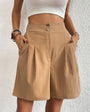 Model wearing khaki high-waisted pleated shorts with white crop top and layered bracelets against textured white wall