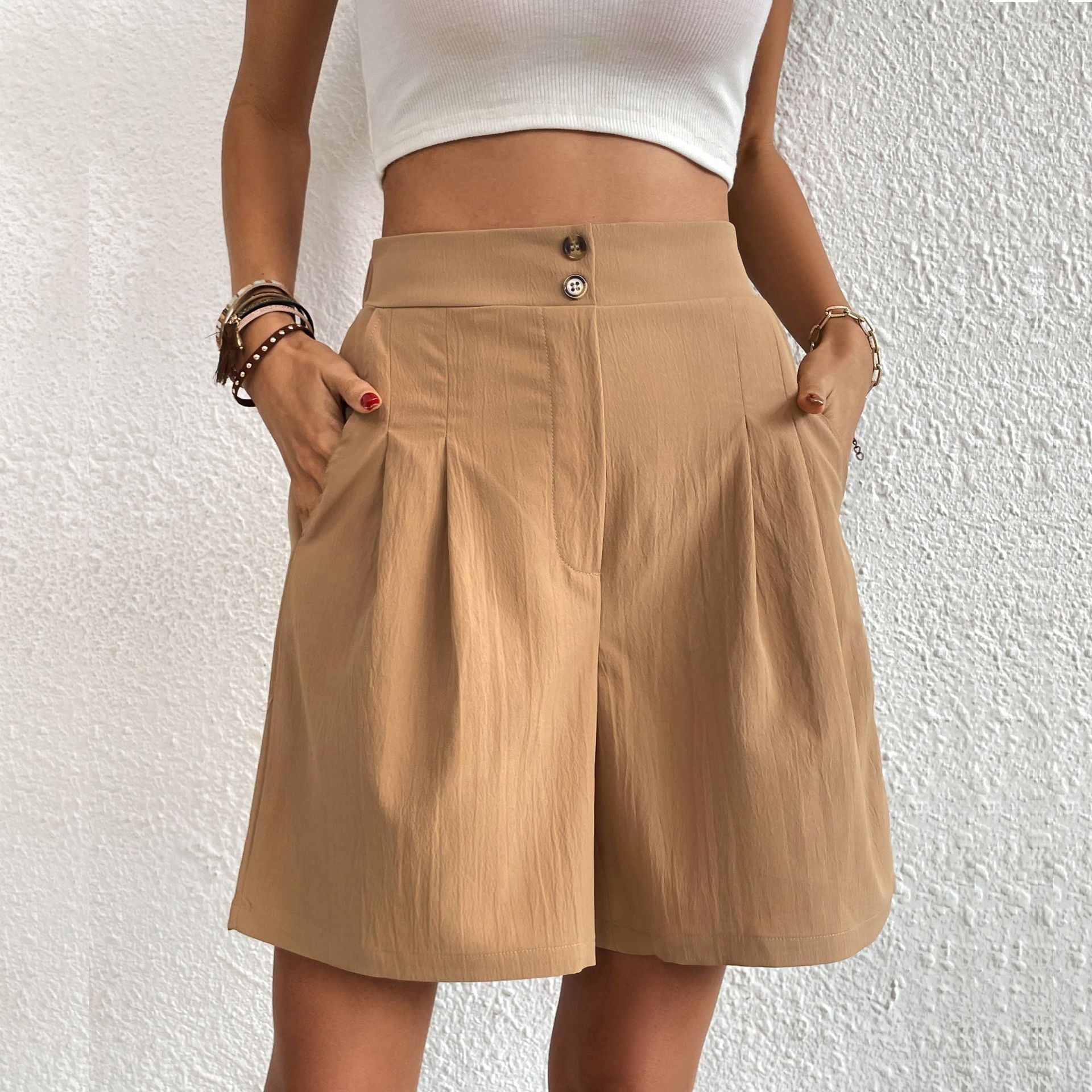 Model wearing khaki high-waisted pleated shorts with white crop top and layered bracelets against textured white wall