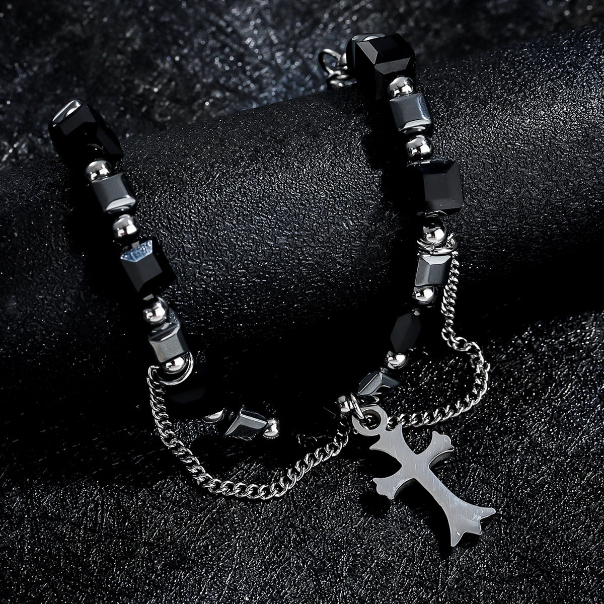 A detailed shot of a black faceted bead bracelet with silver chain accents and a cross pendant, elegantly draped over a black leather surface.