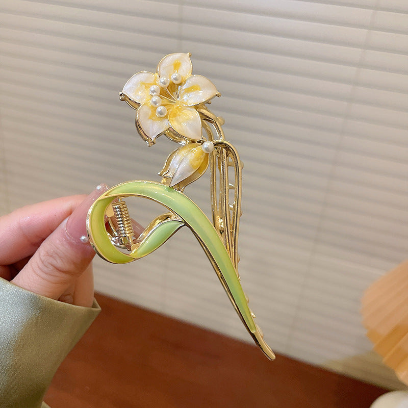 A gold-tone hair claw clip with a yellow enamel flower design, pearl accents, and a curved green leaf motif.