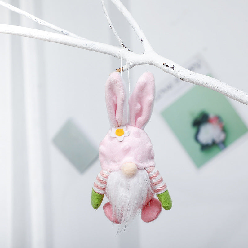 A close-up of a pink plush bunny gnome ornament with a flower detail, hanging from a white tree branch, featuring a fluffy beard.