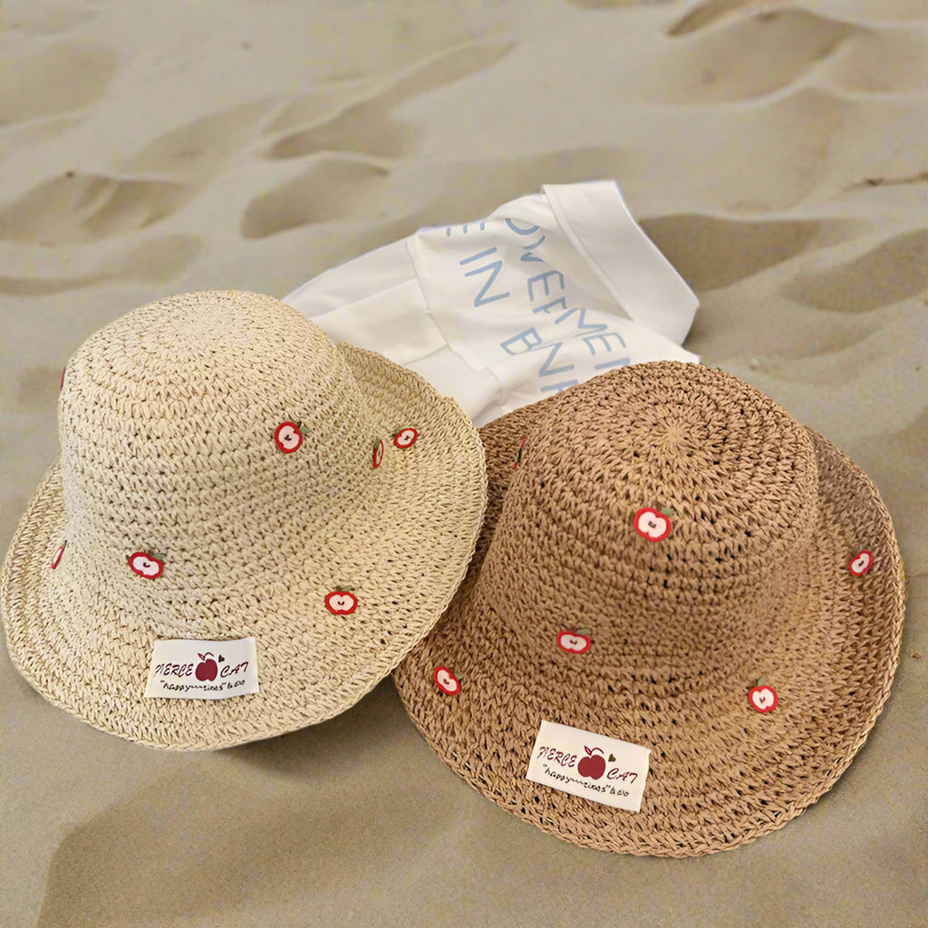 Two woven straw bucket hats with embroidered apple details, one beige and one brown, in a beach setting.