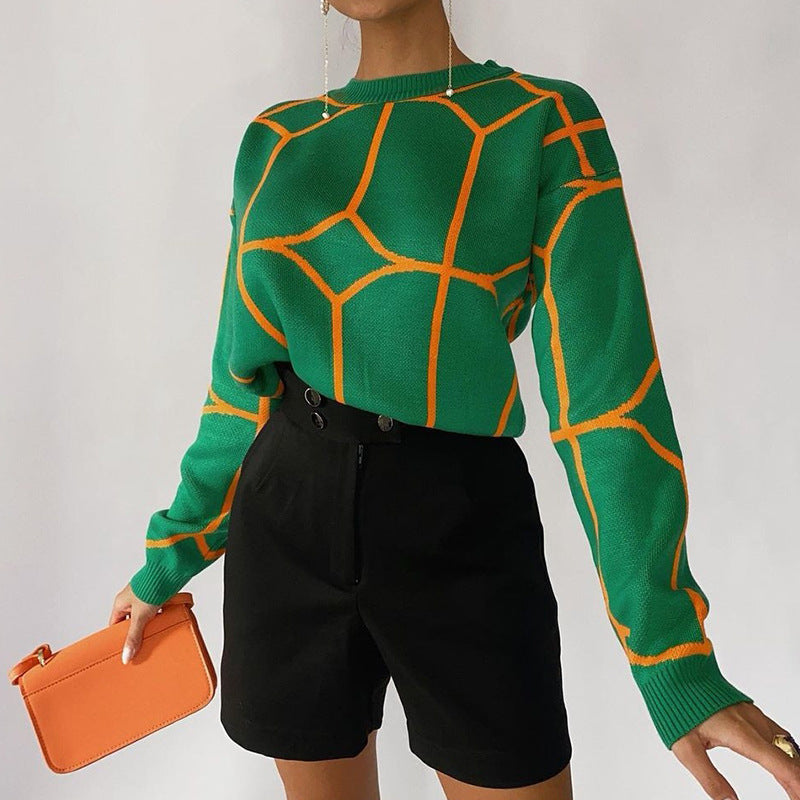A model is standing holding an orange clutch, wearing a green sweater with orange geometric lines and black shorts against white background