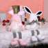 Two seated plush bunny gnome dolls in white and pink, wearing houndstooth-patterned pants, displayed on a fluffy white surface with red baubles.