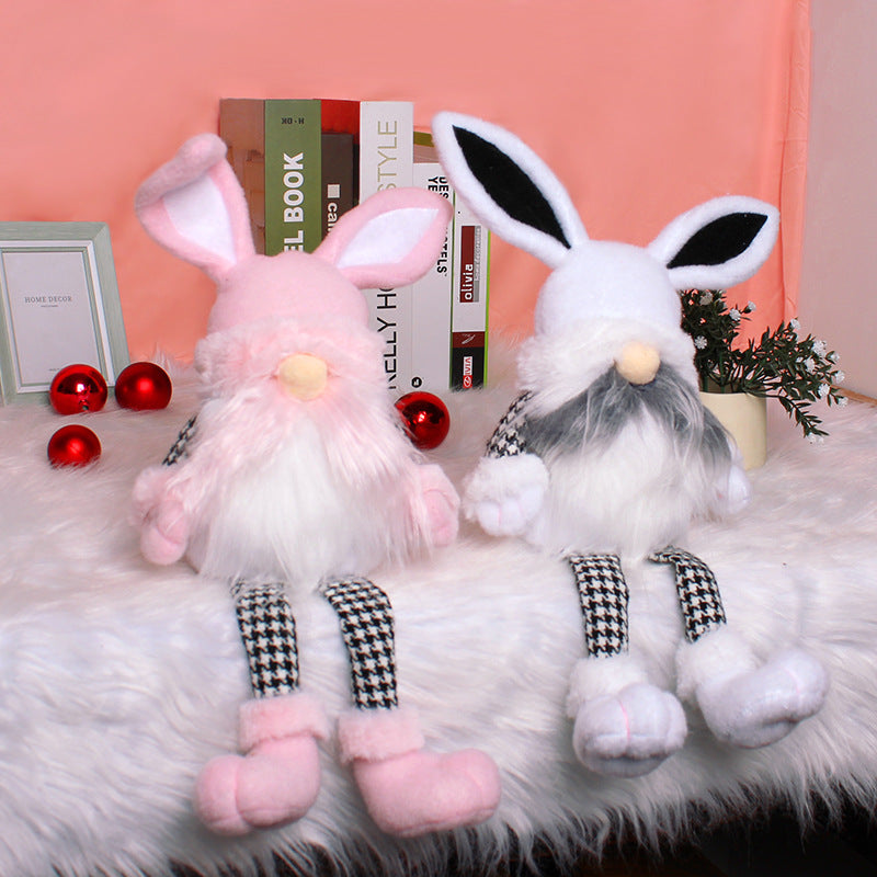 Two seated plush bunny gnome dolls in white and pink, wearing houndstooth-patterned pants, displayed on a fluffy white surface with red baubles.