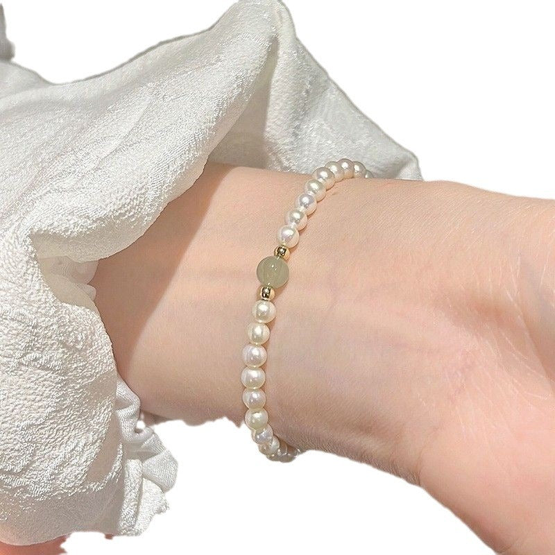 Minimalist bracelet with white pearls and a single light green jade bead, worn against a white textured background