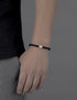 A person wearing a black braided bracelet with a silver charm, with their arm relaxed against a gray background.