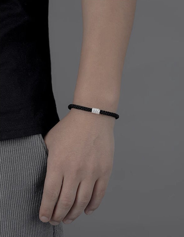 A person wearing a black braided bracelet with a silver charm, with their arm relaxed against a gray background.