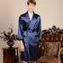 Man in solid navy satin robe with contrast piping detail. Close-up shows patch pockets and belted waist alongside antique nightstand with floral arrangement