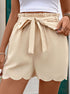 A close-up of beige tie-waist shorts showing paperbag waistband detail and scalloped hem design