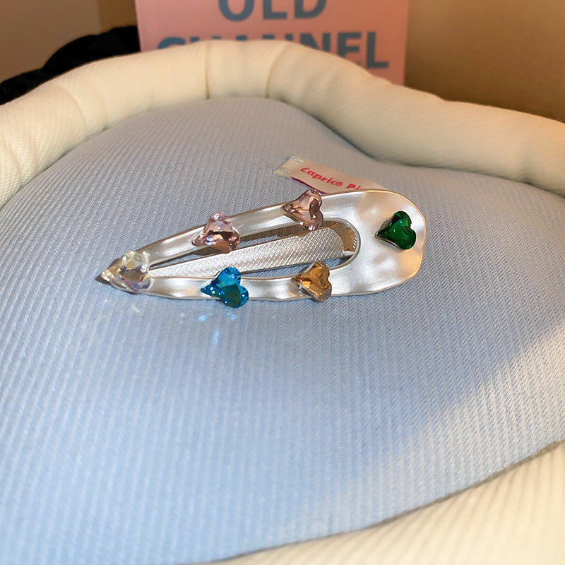 Teardrop-shaped silver hair clip with iridescent gemstones in heart shapes, featuring a pearl accent, displayed on blue fabric.