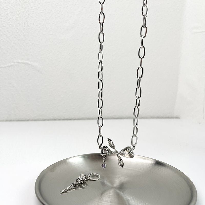 Silver chain necklace with bow charm and key pendant displayed on metal plate. Features oval links and purple gemstone