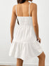 A woman is showing the back view of a white cotton sundress with smocked bodice and adjustable straps, standing against a white wall