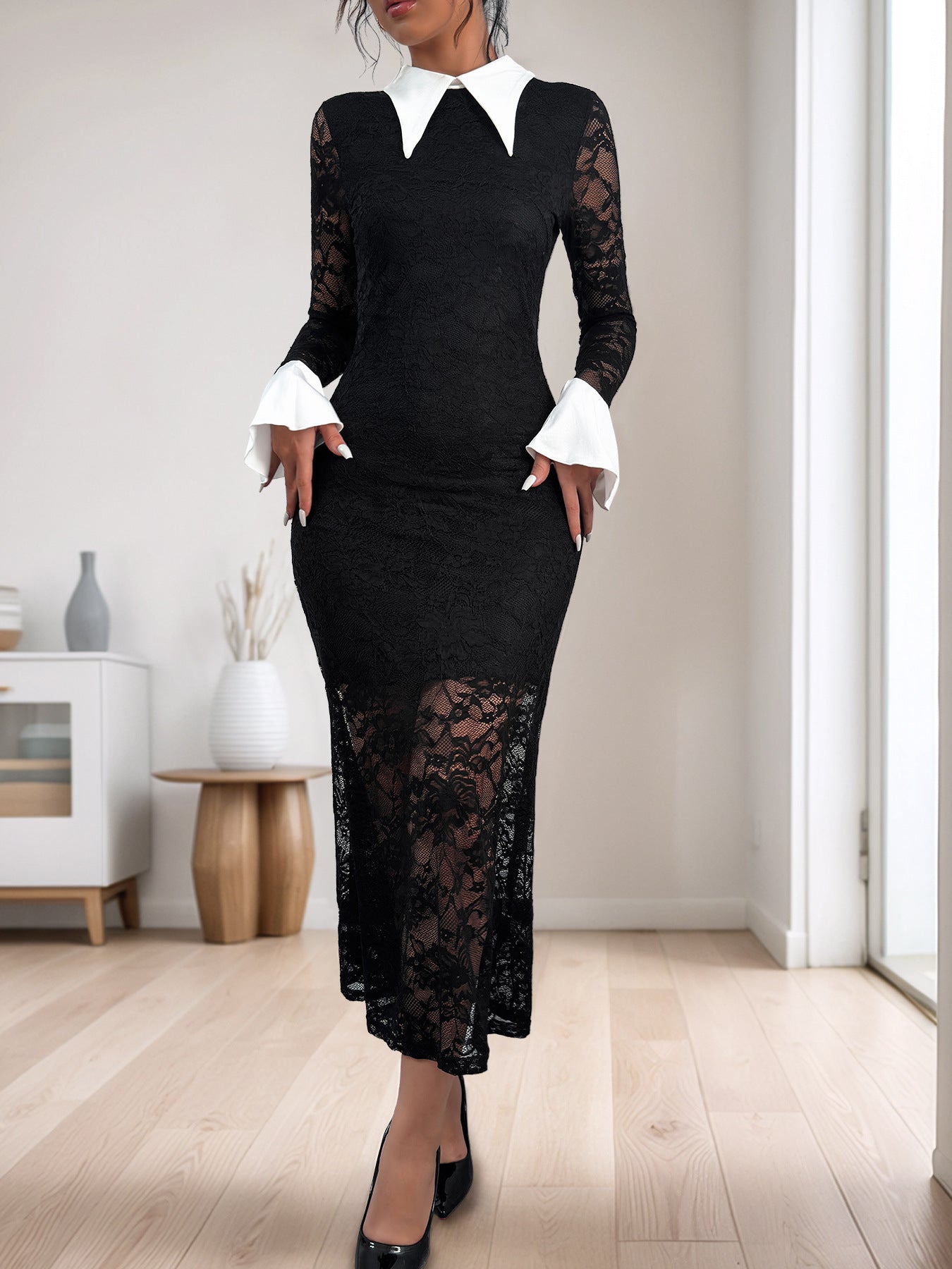 A woman in a fitted black lace dress with sheer lace sleeves, a white pointed collar, and trumpet cuffs, standing in a minimalist room.