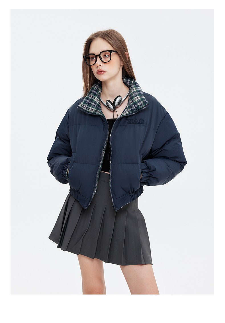 A woman poses in a navy puffer jacket with a plaid lining, a black crop top, a gray pleated skirt, and brown platform boots