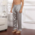Model wearing silver-gray silk pajama pants with repeating circular pattern and white trim, standing barefoot on wooden floor beside white furniture