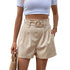 Model wearing beige pleated shorts with circular belt and white crop top, accessorized with straw bag